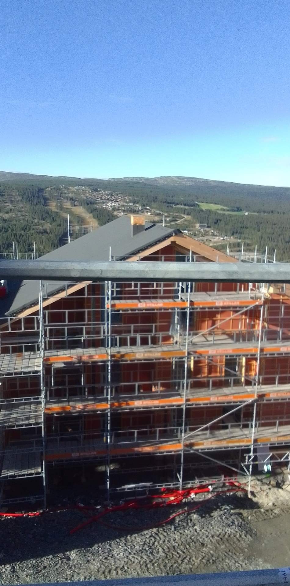 Wznoszony dom z drewnianą konstrukcją szkieletową, widoczny z rusztowania na tle zalesionych wzgórz i błękitnego nieba.