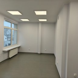 Mateusz Ślepecki - Jasne, odnowione wnętrze gabinetu lekarskiego z wykładziną, oknem i oświetleniem LED. Ściany pomalowane na jasny odcień szarości. Widoczne gniazdka elektryczne.