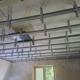 Mateusz Ślepecki - Metalowa konstrukcja sufitu podwieszanego z widocznymi profilami i elementami mocującymi, częściowo zakryta płyta. W tle okno i surowe ściany.