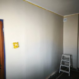 Mateusz Ślepecki - Świeżo pomalowane wnętrze z zabezpieczonymi listwami sufitowymi taśmą malarską, drewnianymi drzwiami z niebieską folią ochronną i aluminiową drabiną.