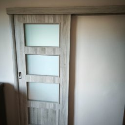 Mateusz Ślepecki - Zamontowane przesuwne drzwi z szarego drewna z trzema matowymi szybami, widoczne na tle jasnej ściany i drewnianej podłogi. Minimalistyczny design.
