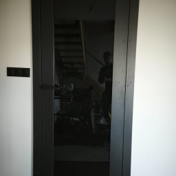 Mateusz Ślepecki - Ciemne, nowoczesne drzwi z czarnym szkłem, zamontowane w białej ścianie. Widoczne odbicie wnętrza z schodami i krzesłem. Minimalistyczny design i precyzyjny montaż.