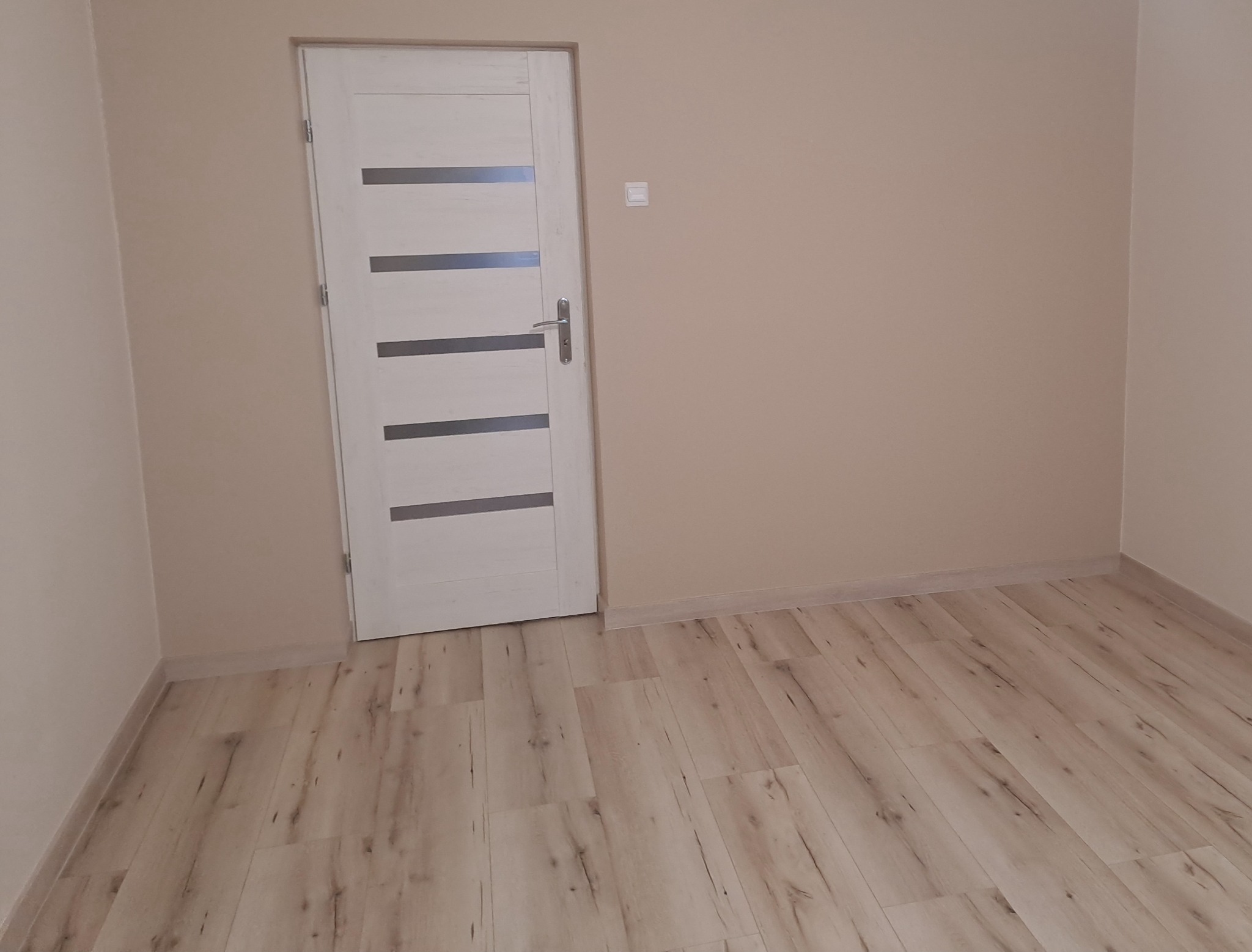 Wnętrze pokoju po remoncie: białe drzwi z poziomymi, szarymi wstawkami, jasne panele podłogowe imitujące drewno, ściany pomalowane na beżowo, widoczny włącznik światła na ścianie.
