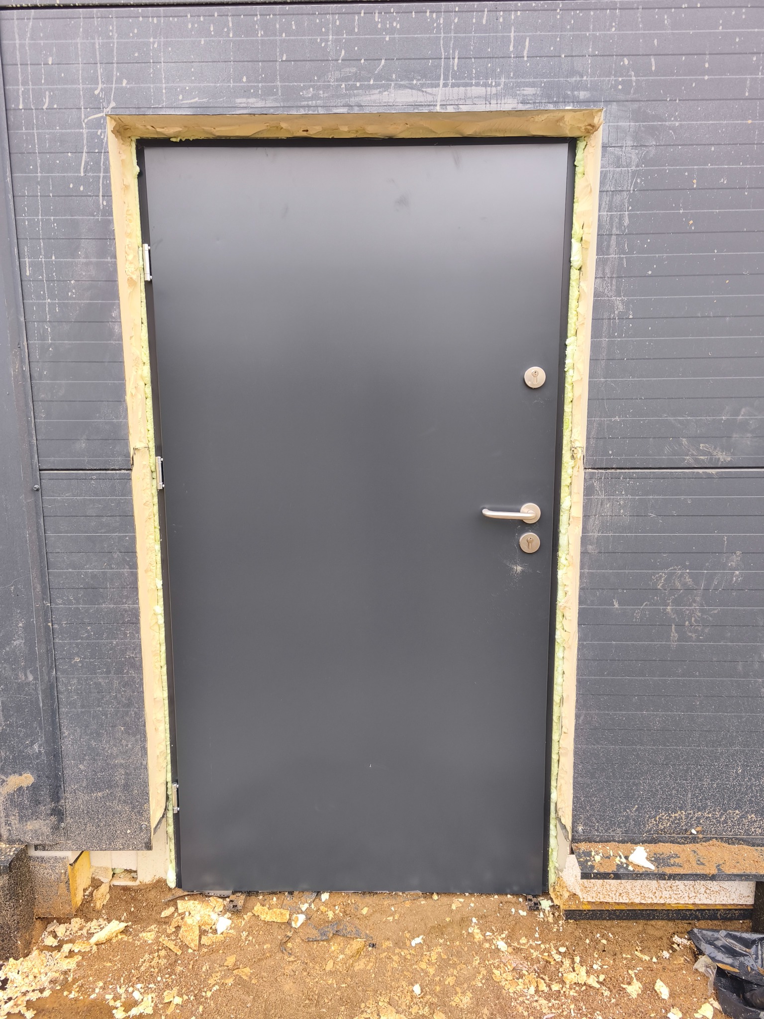 Szare drzwi zewnętrzne, osadzone w ościeżnicy z widoczną pianką montażową, na tle szarej elewacji z poziomymi panelami. Widoczne ślady budowy.