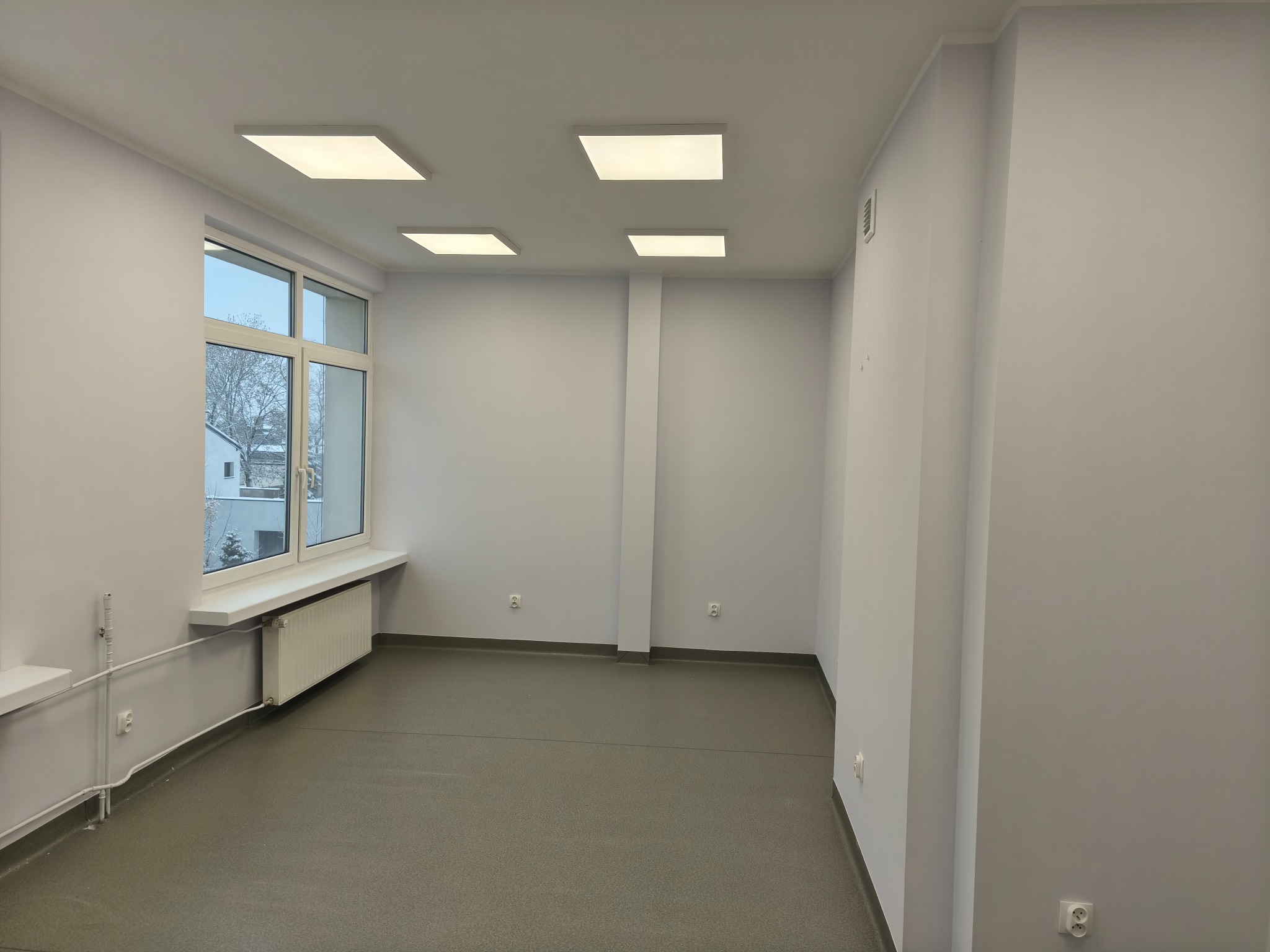 Jasne, odnowione wnętrze gabinetu lekarskiego z wykładziną, oknem i oświetleniem LED. Ściany pomalowane na jasny odcień szarości. Widoczne gniazdka elektryczne.