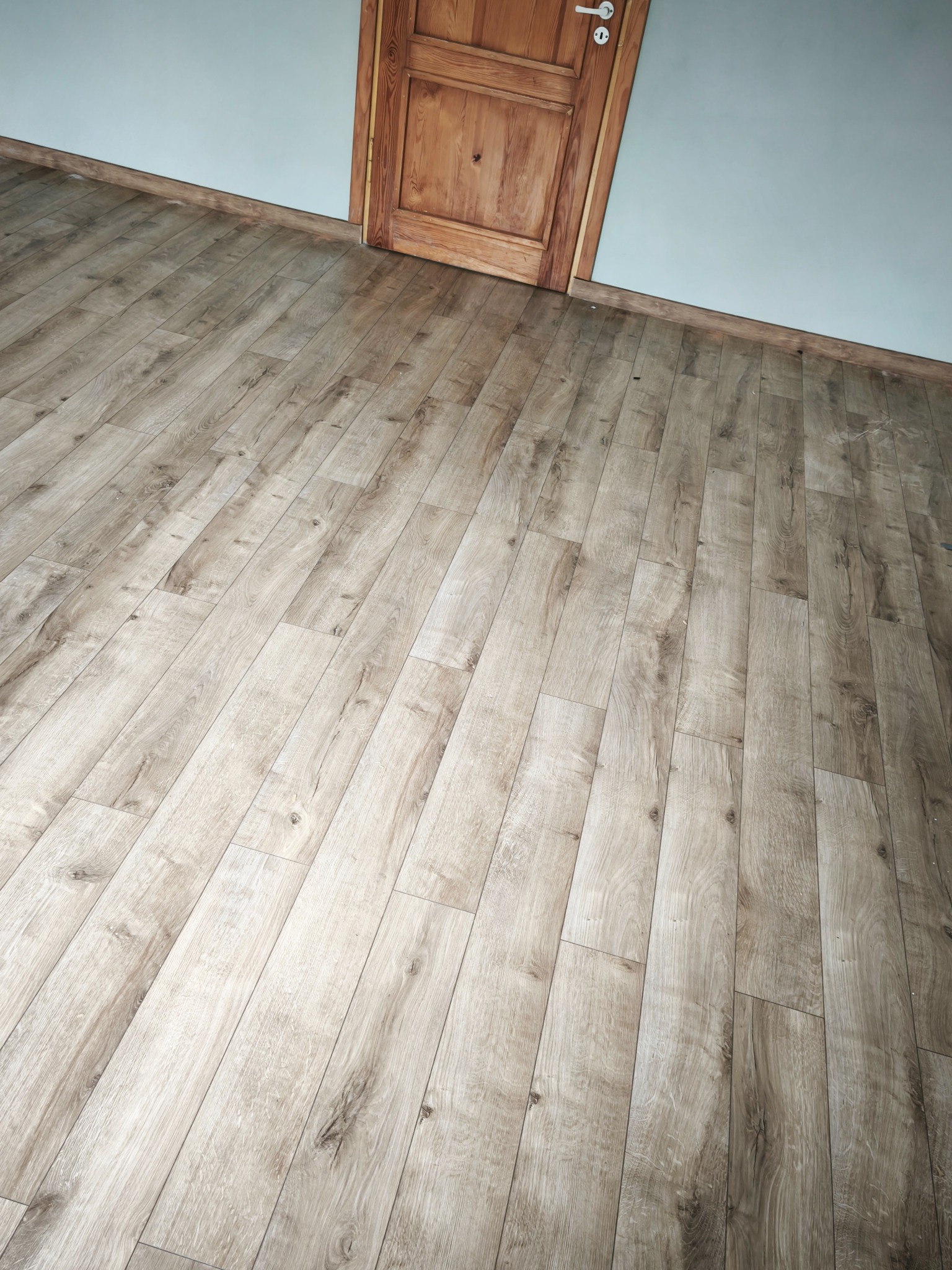 Jasne panele podłogowe ułożone pod kątem, drewniane drzwi z klamką w tle, ściana w odcieniach szarości. Widok z góry, ukończona podłoga.