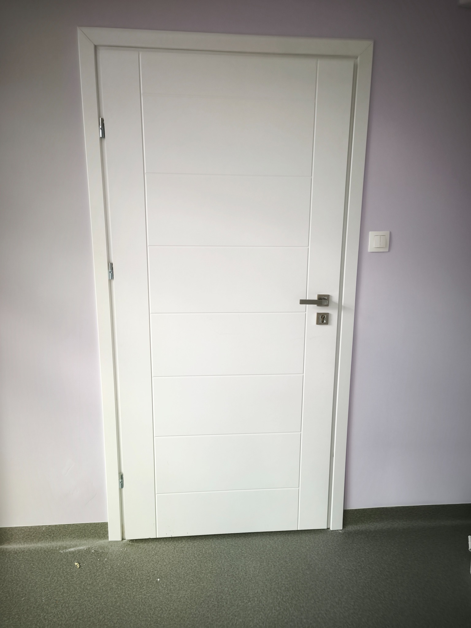Białe, nowoczesne drzwi wewnętrzne z poziomymi frezowaniami i srebrną klamką na tle fioletowej ściany i szarej podłogi. Widoczny włącznik światła.