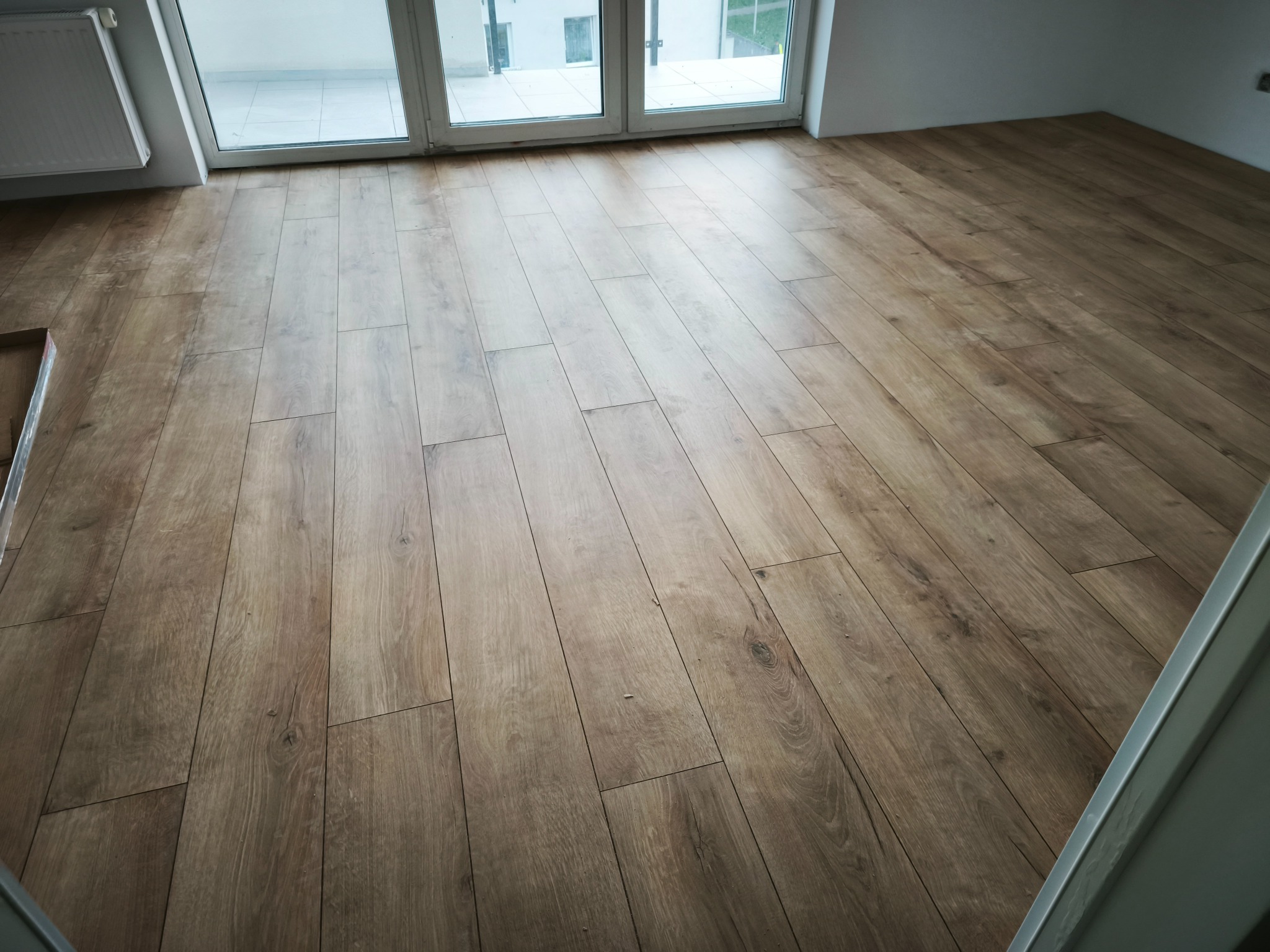 Ułożone panele podłogowe w jasnym odcieniu drewna, widoczne przy oknie balkonowym. Fragment grzejnika i ściany w tle. Widok z góry, ukośny kadr.