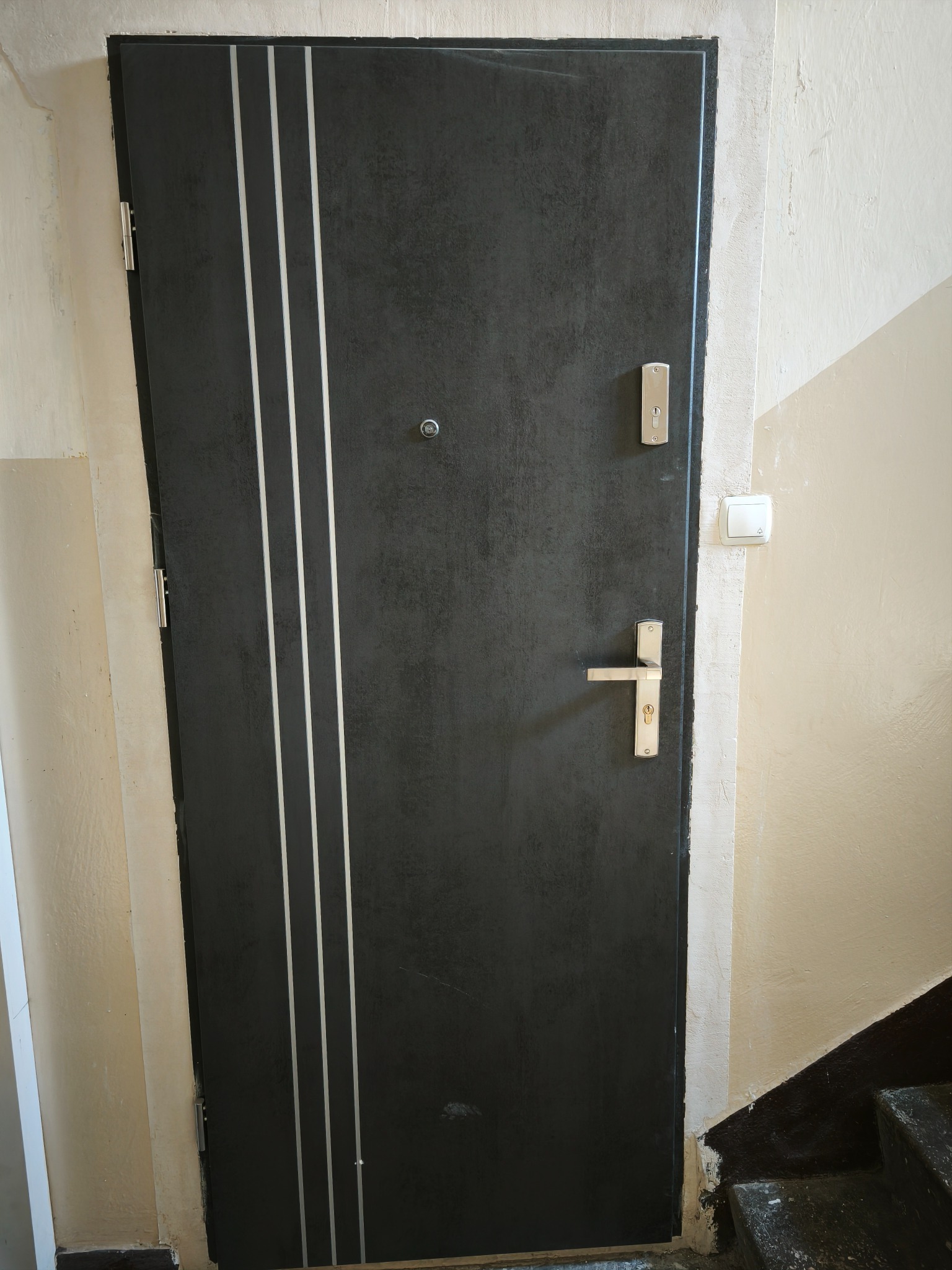 Ciemne drzwi wejściowe z trzema srebrnymi paskami, zamontowane w bloku w Kutnie. Widoczny zamek, klamka i wizjer. Ściana pomalowana na beżowo.