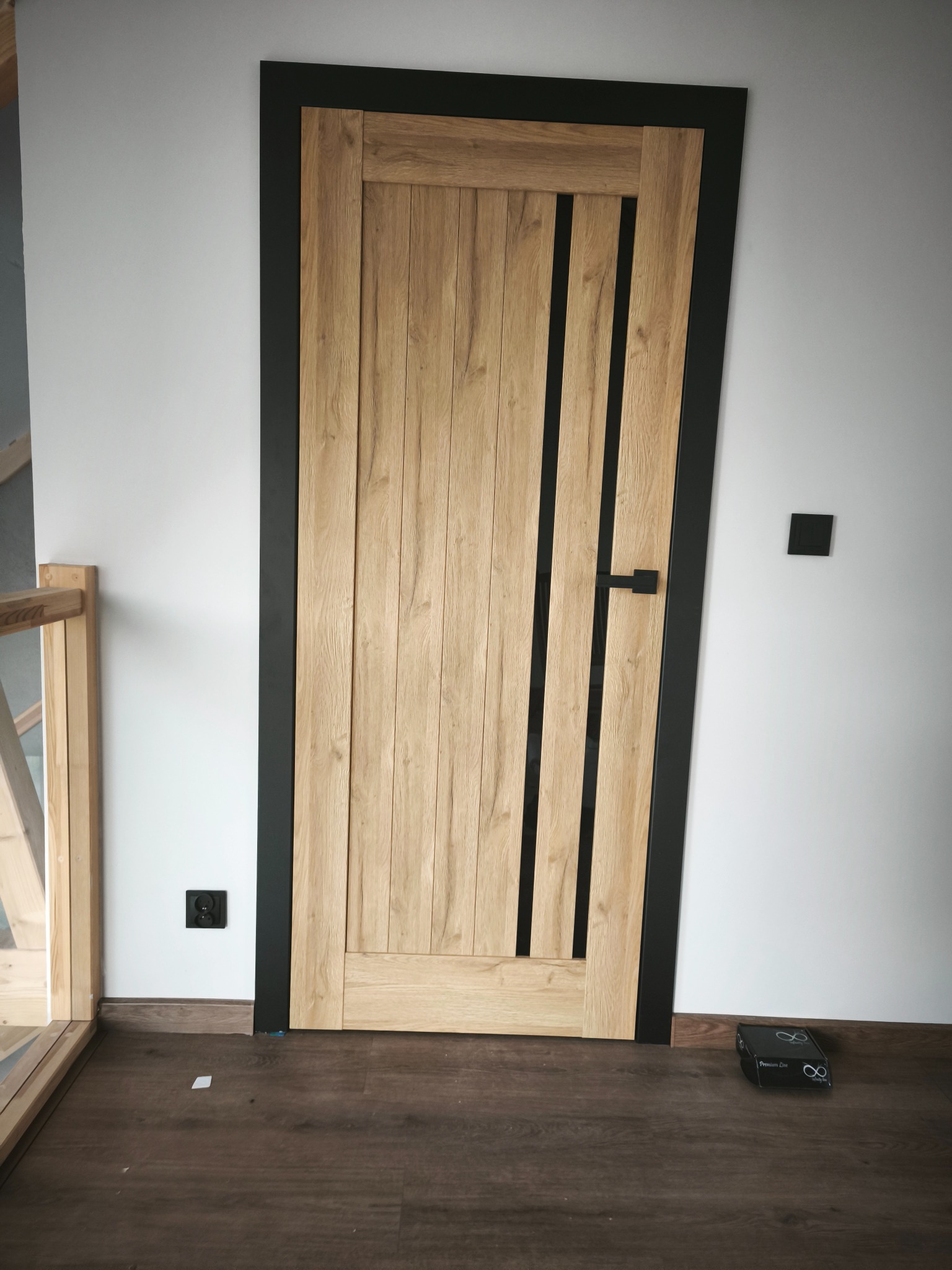 Zamontowane drzwi wewnętrzne z drewnianą okleiną i czarną ościeżnicą, widoczne na tle białej ściany i paneli podłogowych. Minimalistyczny design, nowoczesne wykończenie.