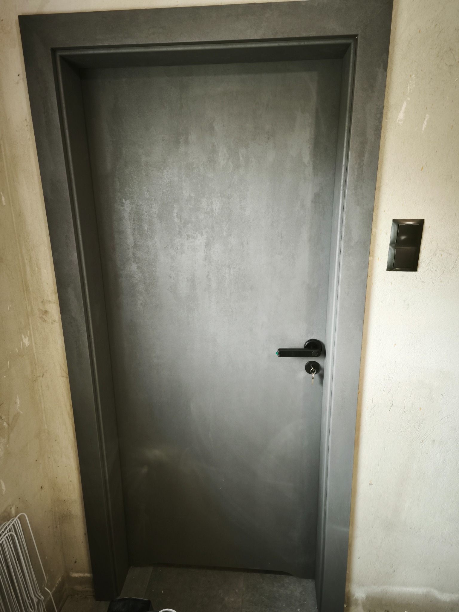 Szare, nowoczesne drzwi wewnętrzne z czarną klamką i kluczem w surowym wnętrzu. Widoczny fragment ściany przed malowaniem i czarny włącznik światła.