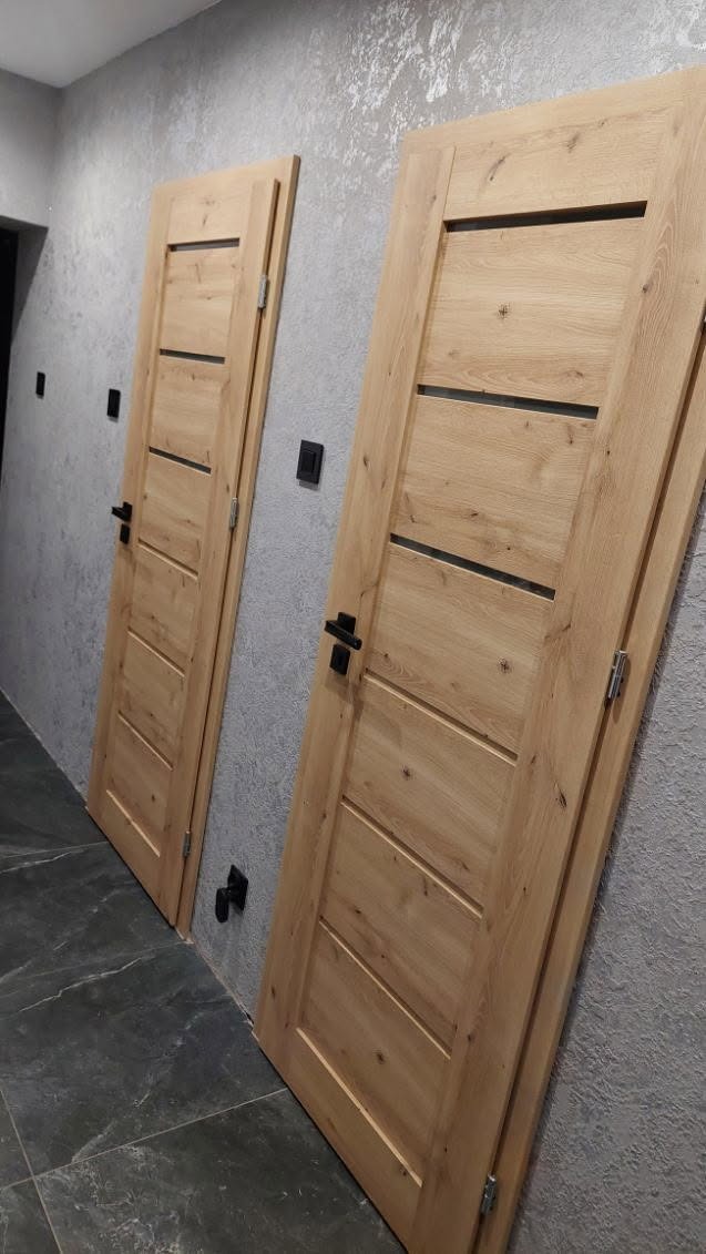 Dwa drewniane drzwi wewnętrzne z czarnymi klamkami, zamontowane w szarej ścianie o dekoracyjnej fakturze. Podłoga wyłożona ciemnymi płytkami imitującymi kamień.