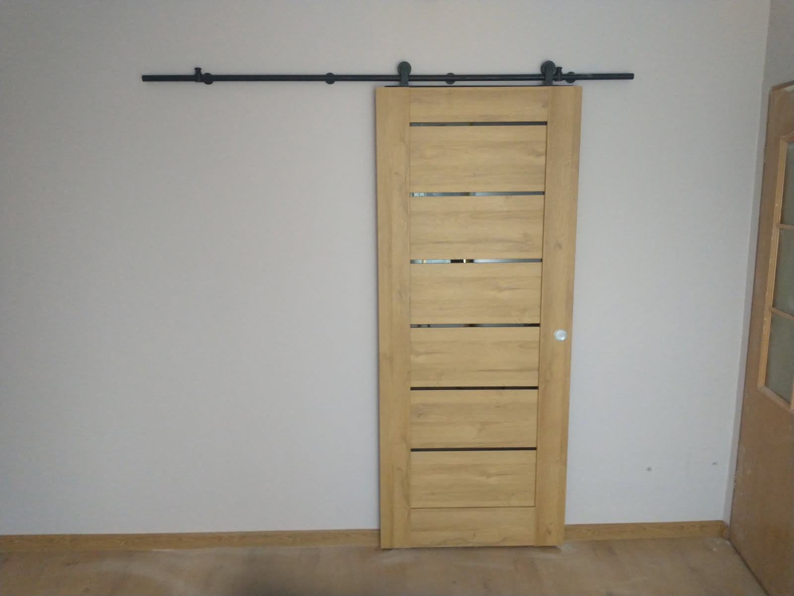 Zamontowane przesuwne drzwi drewniane z czarnymi, poziomymi wstawkami i systemem przesuwnym na szynie. Widoczna ściana i fragment podłogi.