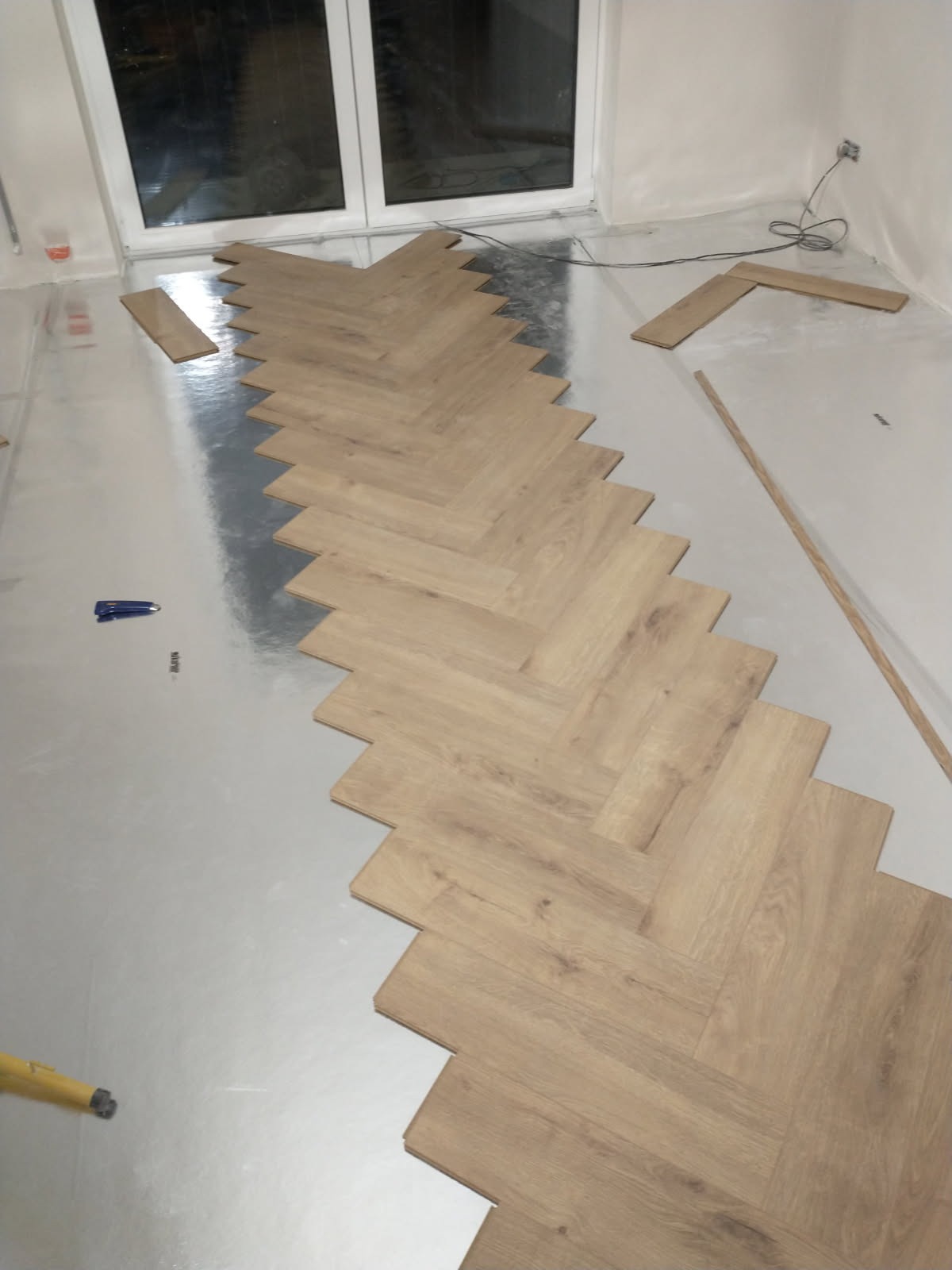 Układanie jasnych paneli podłogowych w jodełkę na srebrnej folii izolacyjnej. Widoczne narzędzia: zszywacz i poziomica. Częściowo ułożona podłoga w nowym pomieszczeniu.