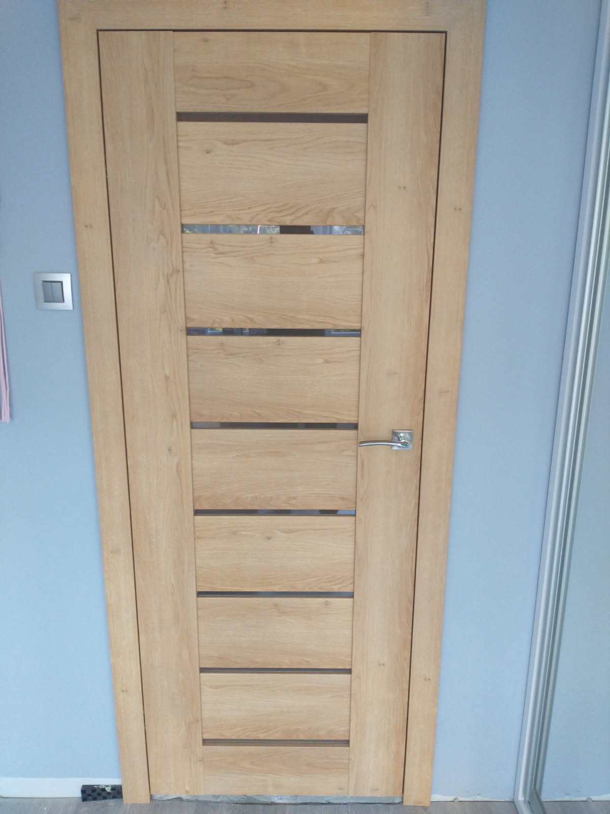 Jasne, drewniane drzwi wewnętrzne z poziomymi przeszkleniami, zamontowane w ścianie o błękitnym odcieniu. Widoczna klamka i fragment listwy przypodłogowej.