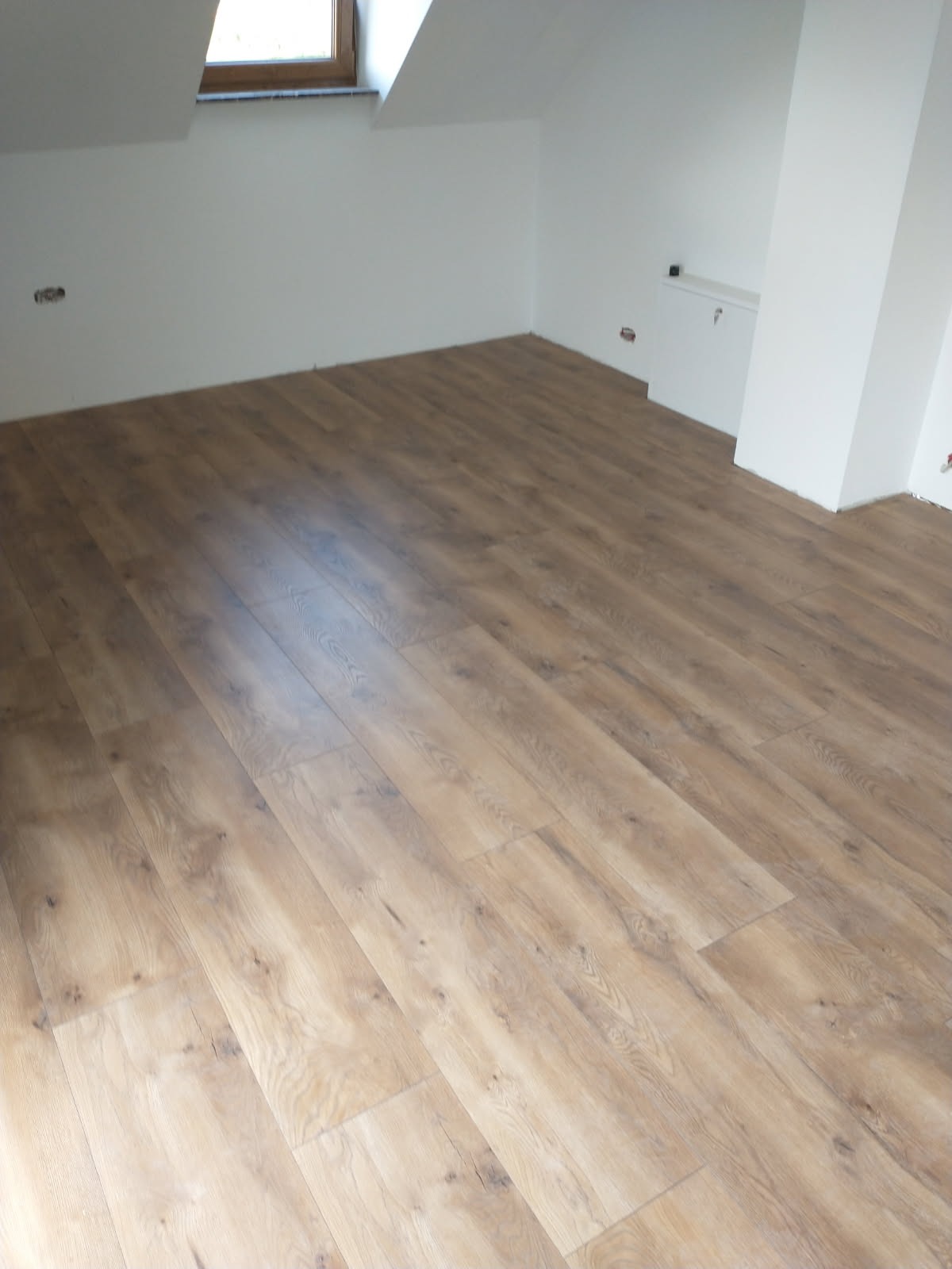 Jasne panele podłogowe ułożone w pokoju na poddaszu z oknem dachowym. Widoczne białe ściany i fragment kolumny. Podłoga z wyraźnym wzorem drewna.