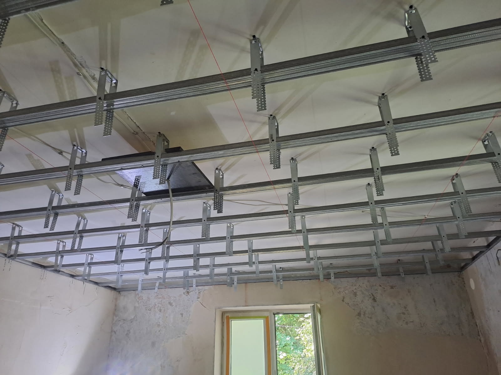Metalowa konstrukcja sufitu podwieszanego z widocznymi profilami i elementami mocującymi, częściowo zakryta płyta. W tle okno i surowe ściany.