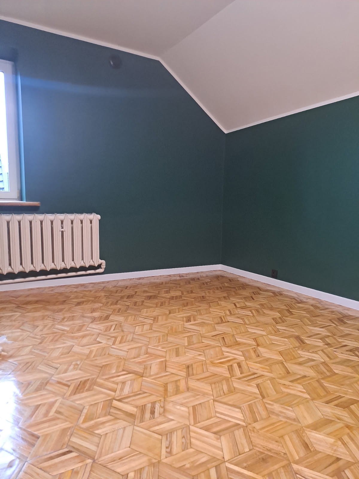 Pokój po remoncie: parkiet w jodełkę, ściany pomalowane na ciemnozielono, białe listwy przypodłogowe, grzejnik pod oknem. Widok na narożnik pokoju.
