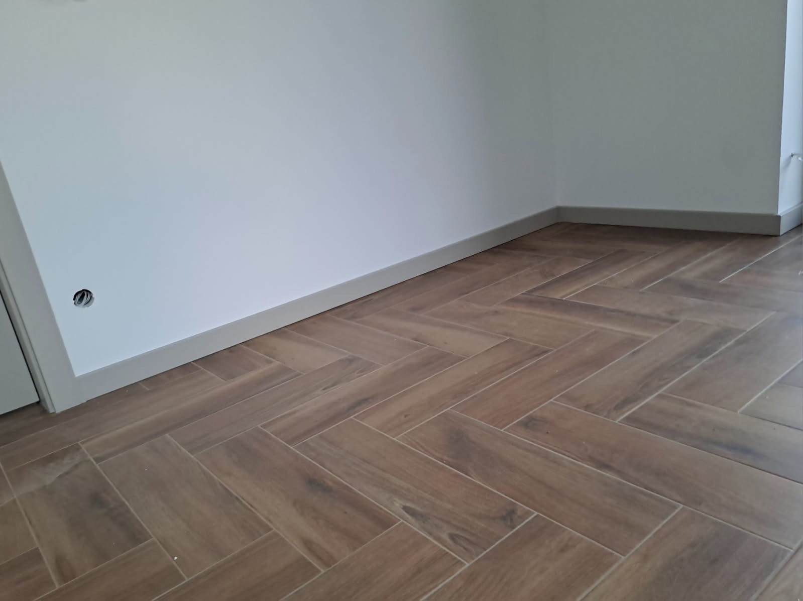 Ułożona podłoga z płytek imitujących drewno w jodełkę, szara listwa przypodłogowa, białe ściany z gniazdkiem. Widok na róg pokoju.