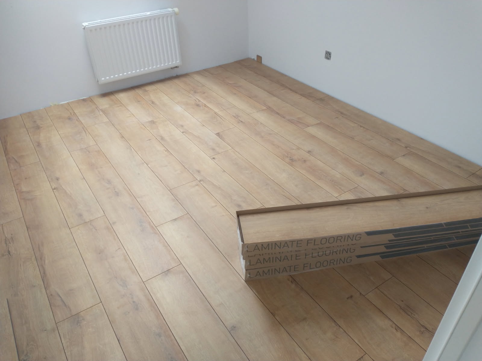 Jasne panele podłogowe ułożone w pokoju, widoczny grzejnik na ścianie i paczka paneli. Częściowo widoczne listwy przypodłogowe.