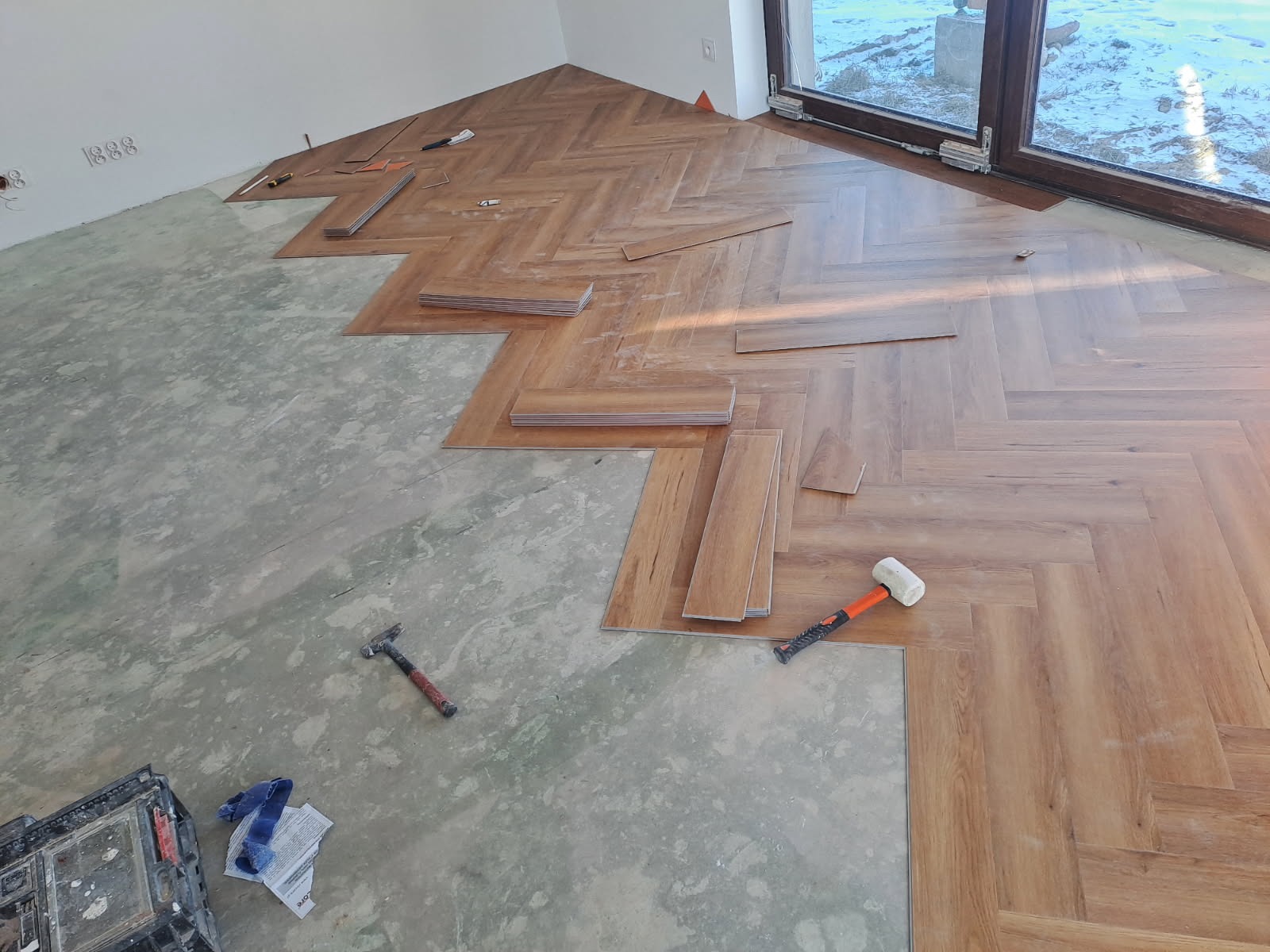 Układanie paneli podłogowych w jodełkę, widoczne narzędzia i częściowo ułożona podłoga. Fragment okna z widokiem na zewnątrz. Jasne wnętrze w trakcie prac wykończeniowych.