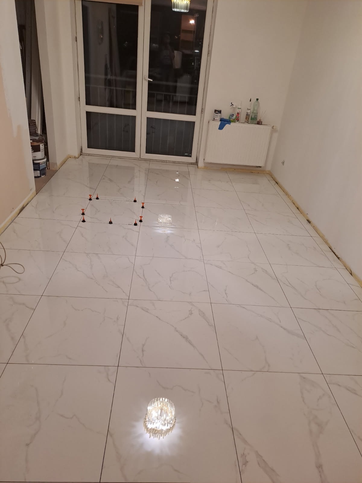 Błyszczące, białe płytki imitujące marmur, ułożone w pokoju z balkonem. Widoczne poziomujące klipsy i kliny. Na ścianie grzejnik i butelki z chemią budowlaną.