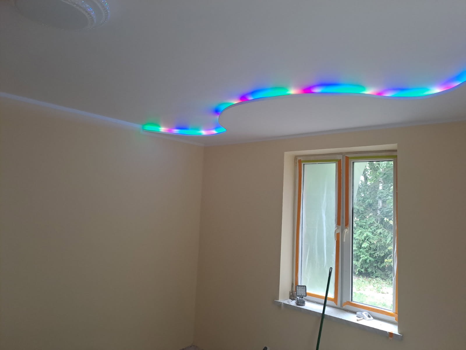 Pomieszczenie po malowaniu z oknem zabezpieczonym folią i sufitem z dekoracyjnym oświetleniem LED w kolorach tęczy. Ściany w odcieniach beżu.