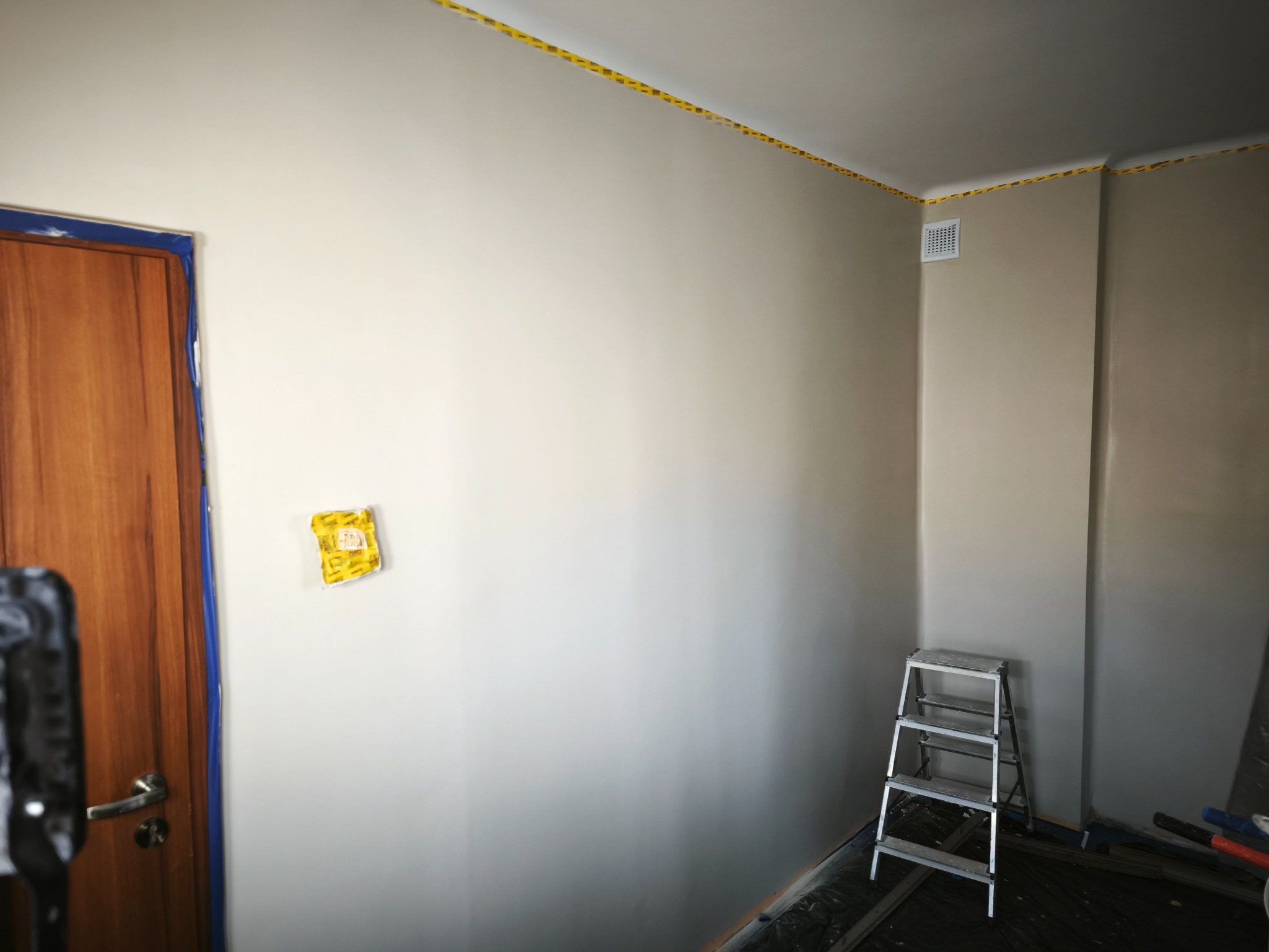 Świeżo pomalowane wnętrze z zabezpieczonymi listwami sufitowymi taśmą malarską, drewnianymi drzwiami z niebieską folią ochronną i aluminiową drabiną.