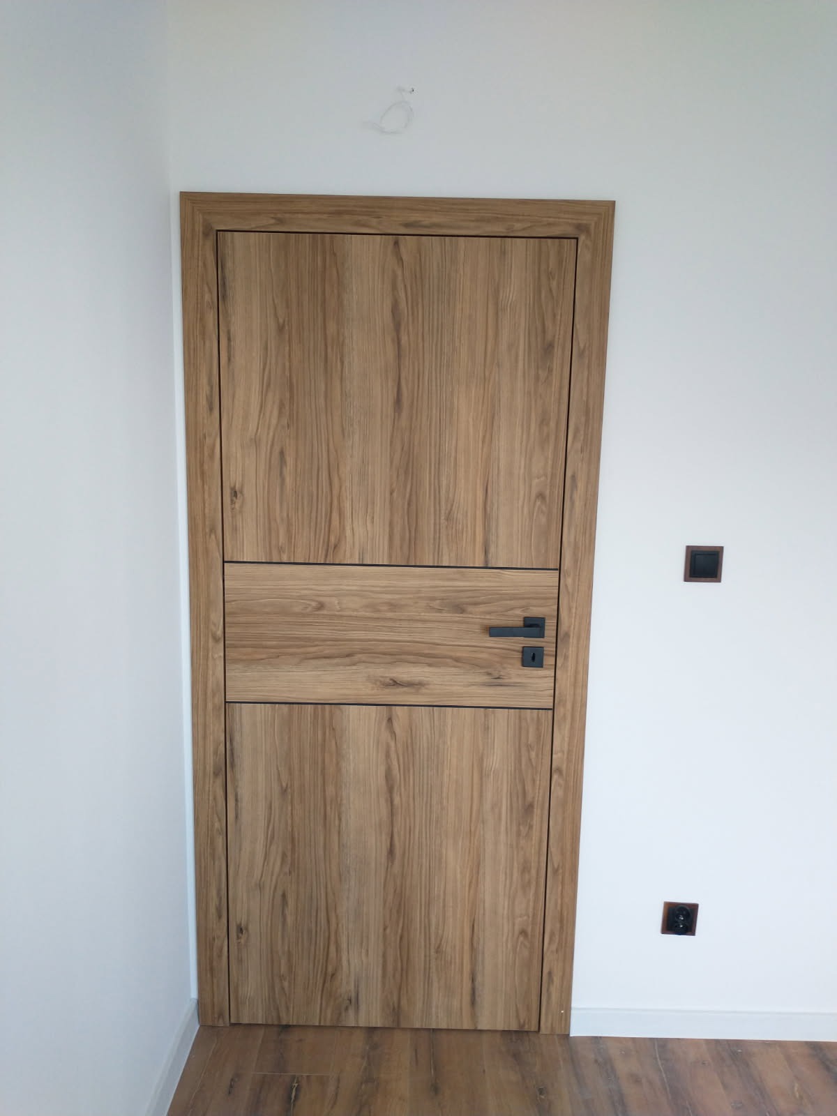 Zamknięte, drewniane drzwi wewnętrzne z czarną klamką, wstawione w białą ścianę z widocznymi gniazdkami elektrycznymi. Podłoga z paneli.