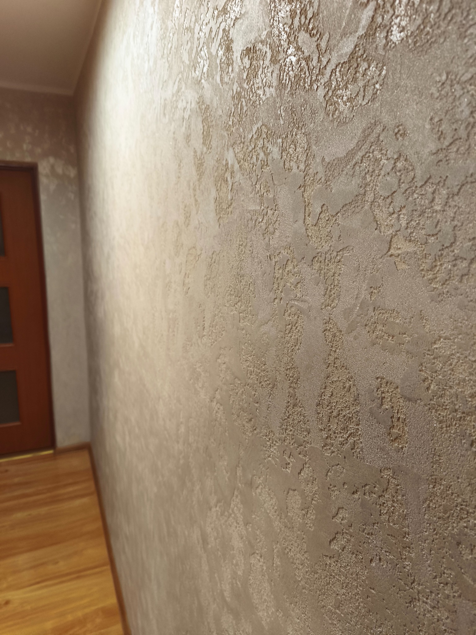 Dekoracyjna ściana z efektem strukturalnym w odcieniach beżu i złota, widoczna z boku, z fragmentem drewnianej podłogi i drzwi w tle. Unikalna faktura ściany.