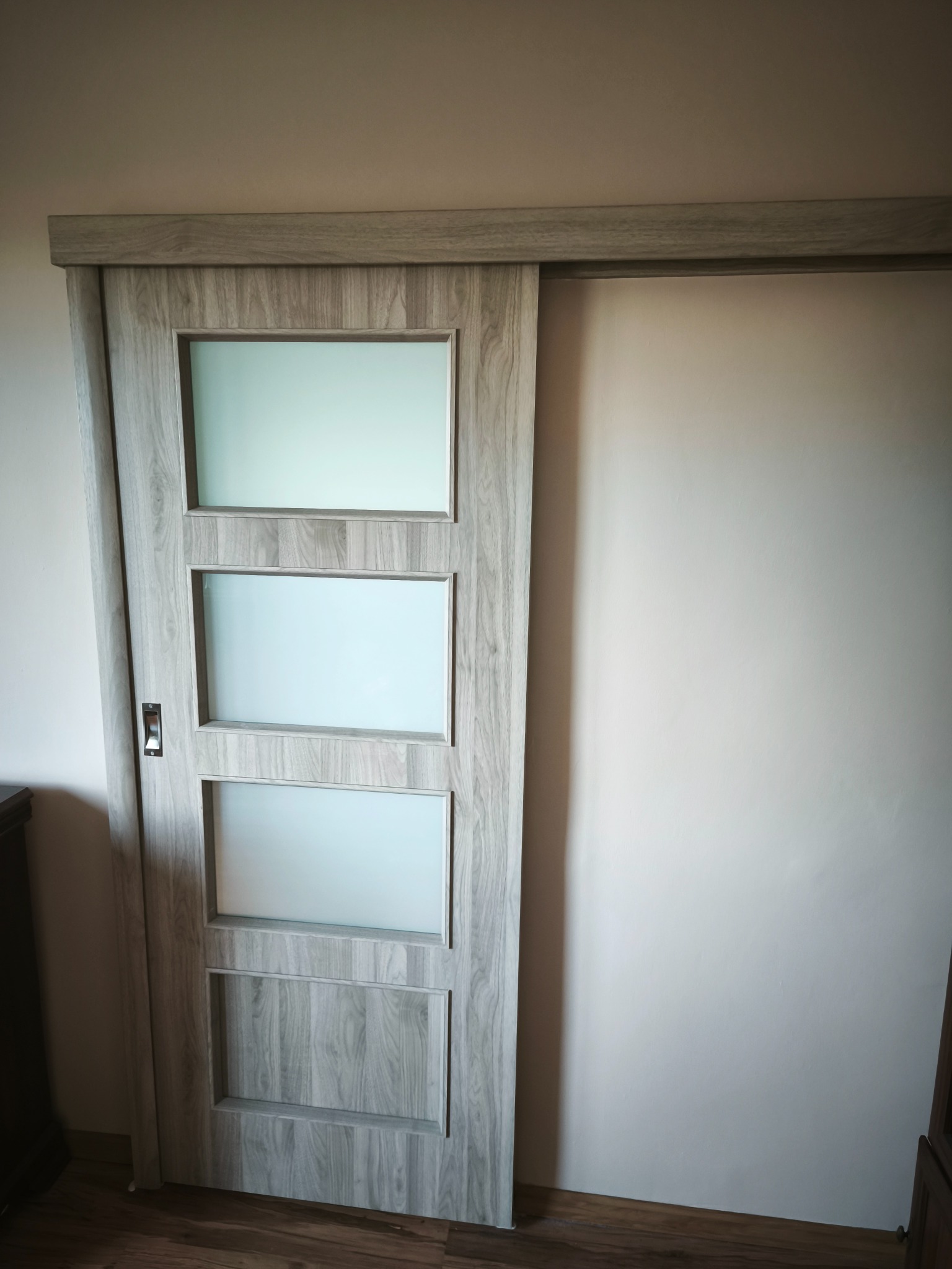 Zamontowane przesuwne drzwi z szarego drewna z trzema matowymi szybami, widoczne na tle jasnej ściany i drewnianej podłogi. Minimalistyczny design.