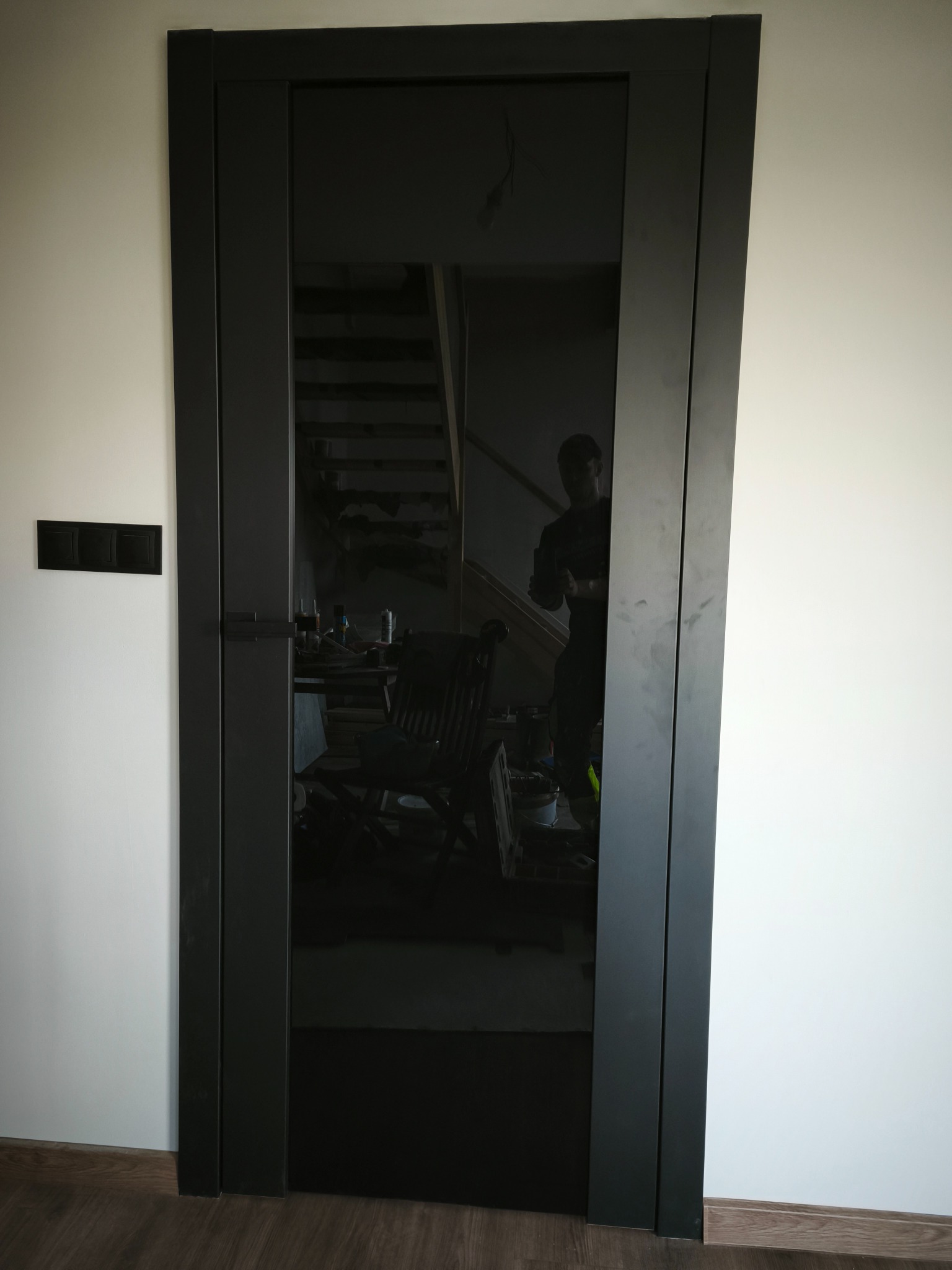 Ciemne, nowoczesne drzwi z czarnym szkłem, zamontowane w białej ścianie. Widoczne odbicie wnętrza z schodami i krzesłem. Minimalistyczny design i precyzyjny montaż.