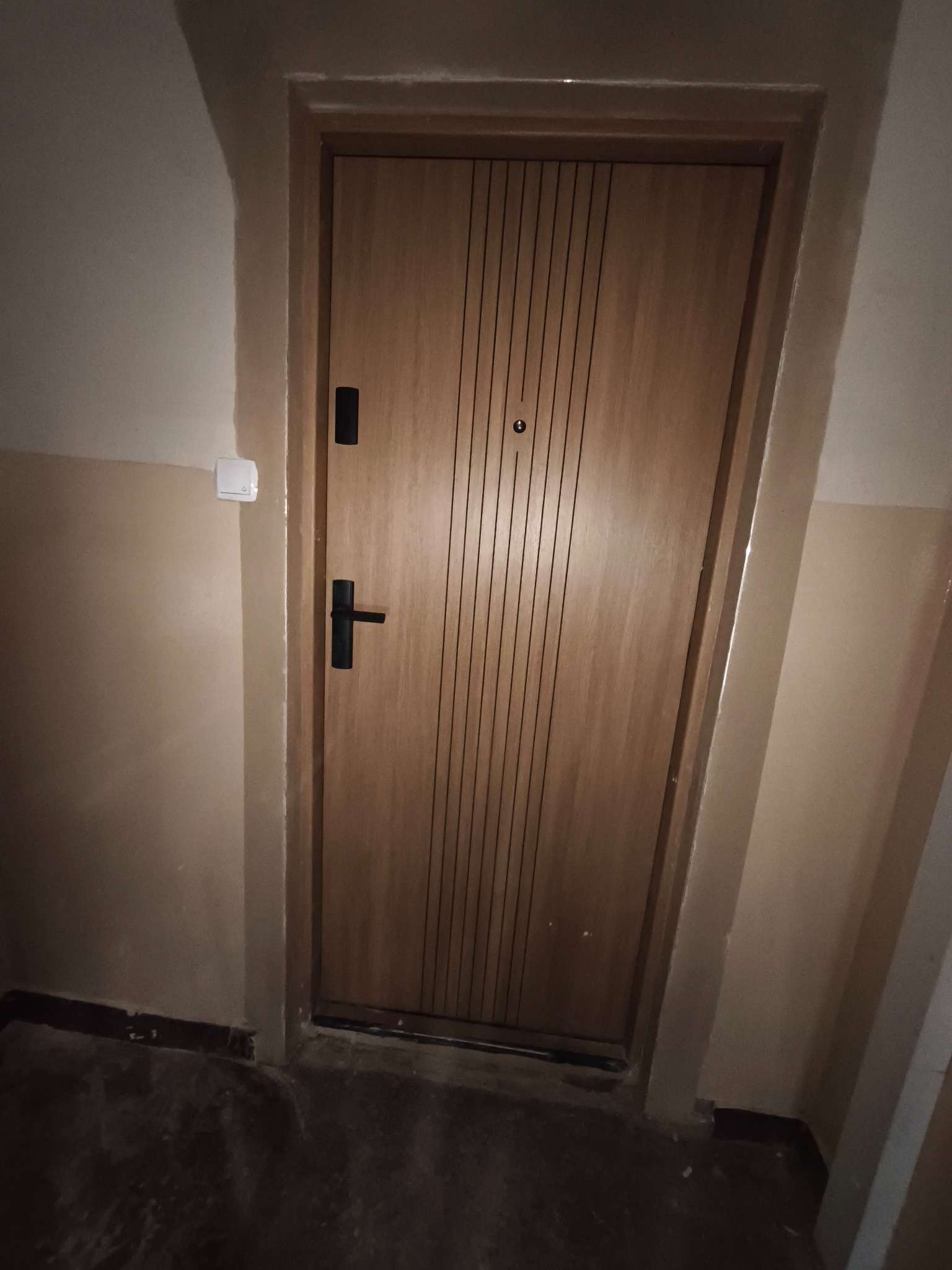 Zamknięte, drewniane drzwi wejściowe z czarną klamką i wizjerem, zamontowane w beżowej ścianie klatki schodowej. Widoczny fragment podłogi i włącznik światła.