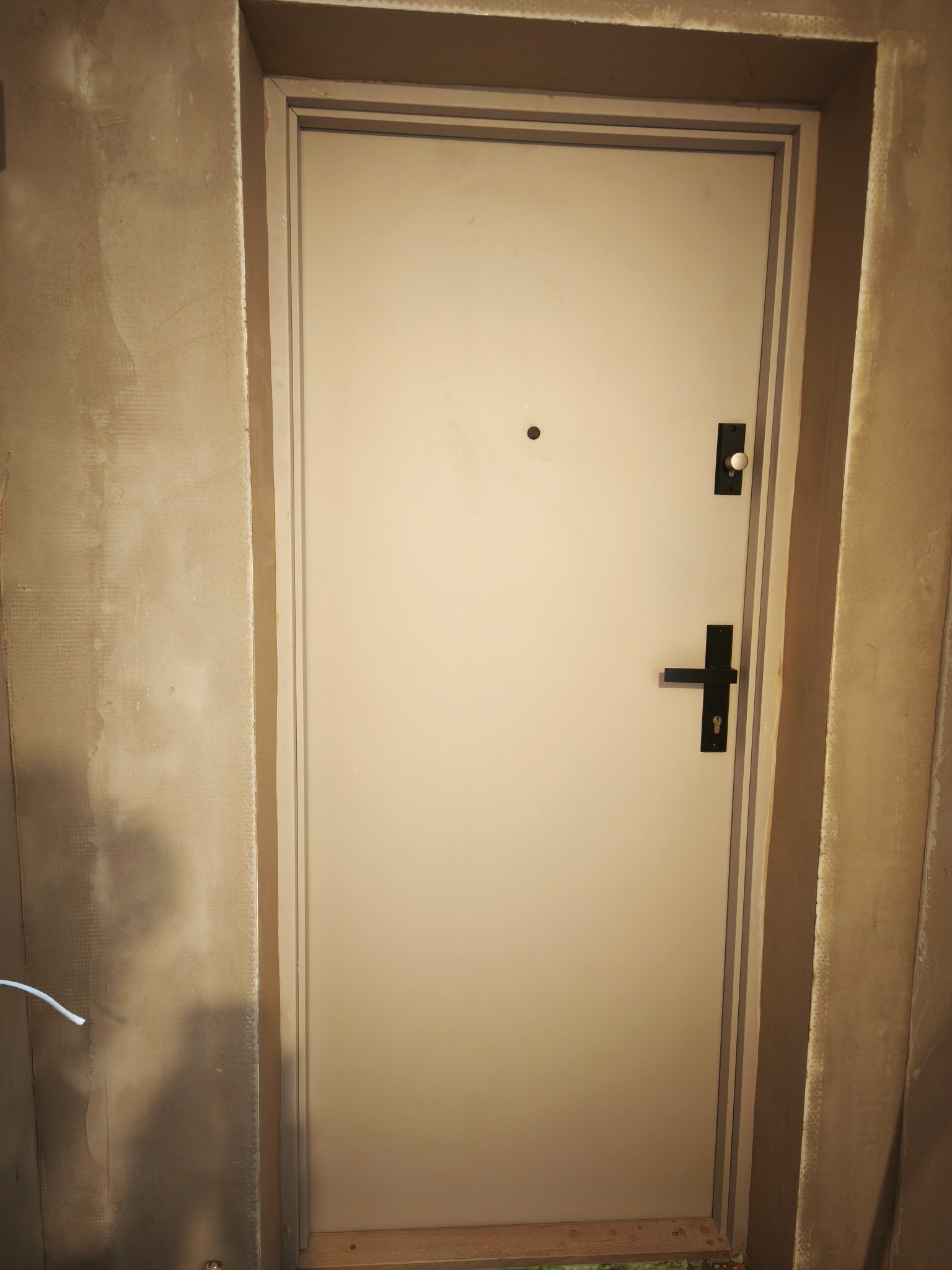 Zamknięte, beżowe drzwi wejściowe z czarną klamką i wizjerem, osadzone w surowym, otynkowanym otworze ściany. Widoczne ślady prac wykończeniowych.