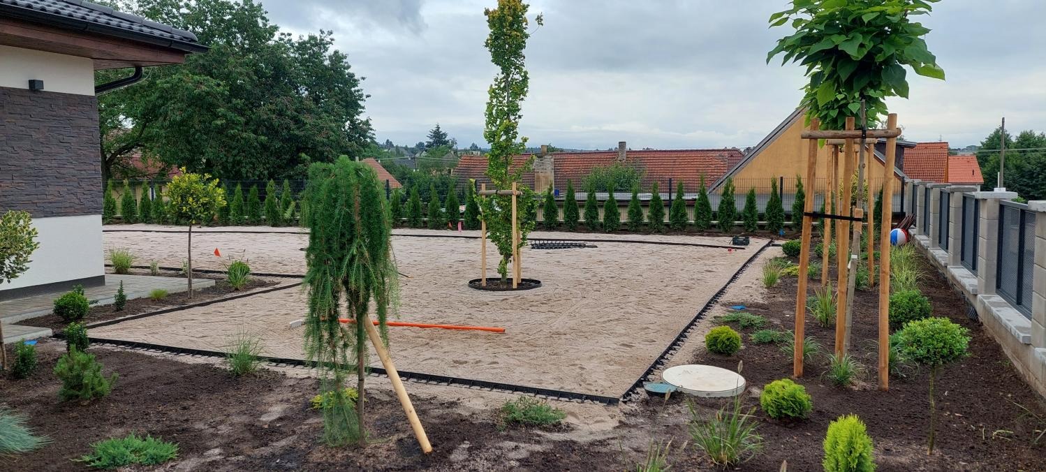 Przygotowany teren pod przyszły ogród z posadzonymi drzewami i krzewami, piaskowa nawierzchnia oddzielona obrzeżami, widoczne podpory drzewek, w tle ogrodzenie.