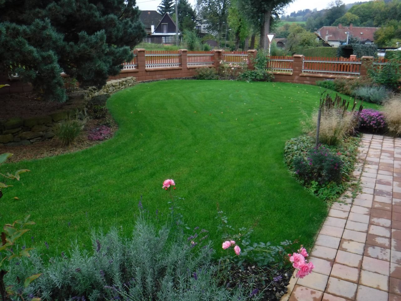 Zadbany trawnik z równo przystrzyżoną trawą, obrzeżony kamiennym murkiem i rabatami kwiatowymi, w tle ogrodzenie z cegły klinkierowej i drewnianych sztachet.