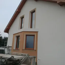 Dom jednorodzinny z oknami w drewnianych ramach, częściowo otynkowany na biało, z widocznymi rusztowaniami i elementami elewacji w odcieniach brązu i beżu.
