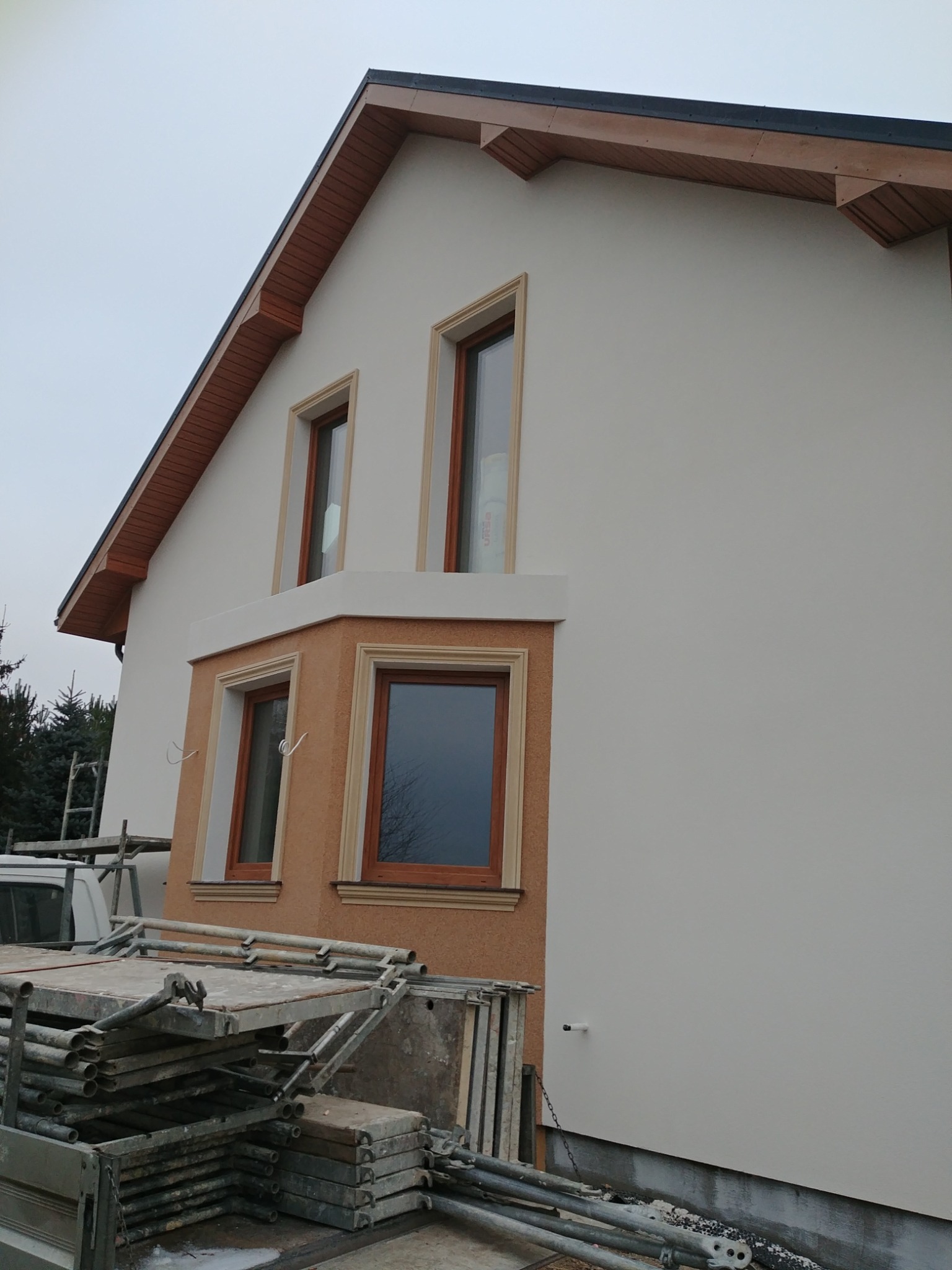 Dom jednorodzinny z oknami w drewnianych ramach, częściowo otynkowany na biało, z widocznymi rusztowaniami i elementami elewacji w odcieniach brązu i beżu.