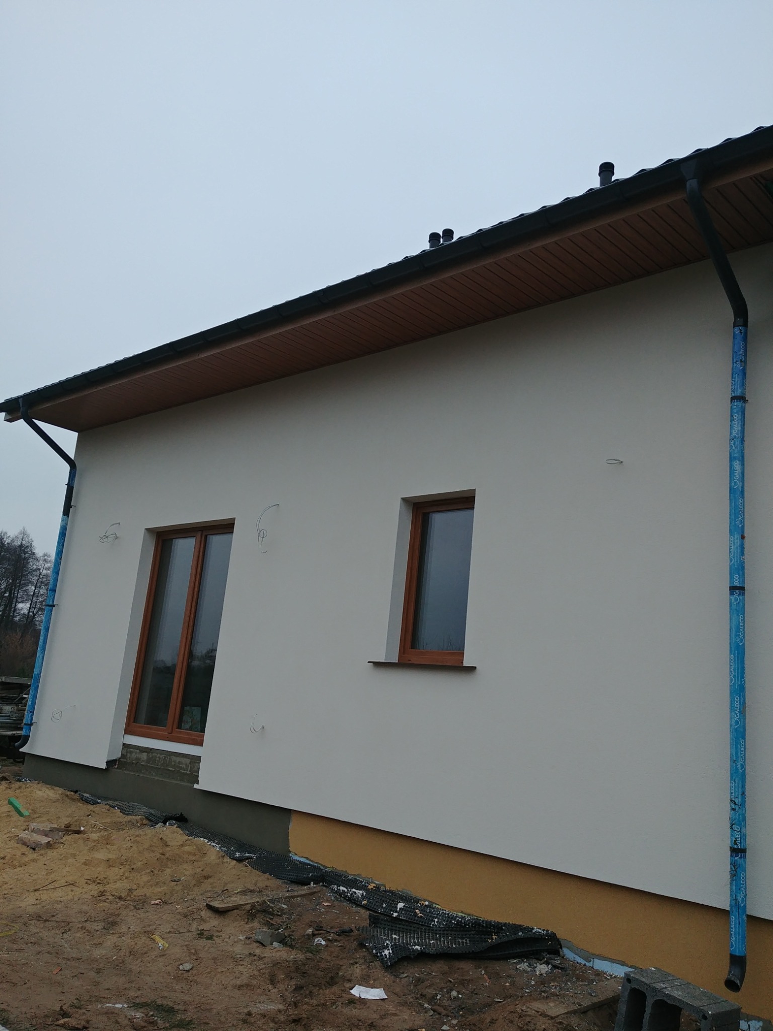 Boczny widok domu z otynkowaną elewacją w trakcie prac wykończeniowych, widoczne okna z brązowymi ramami, orynnowanie i fragment fundamentu z zabezpieczeniem.