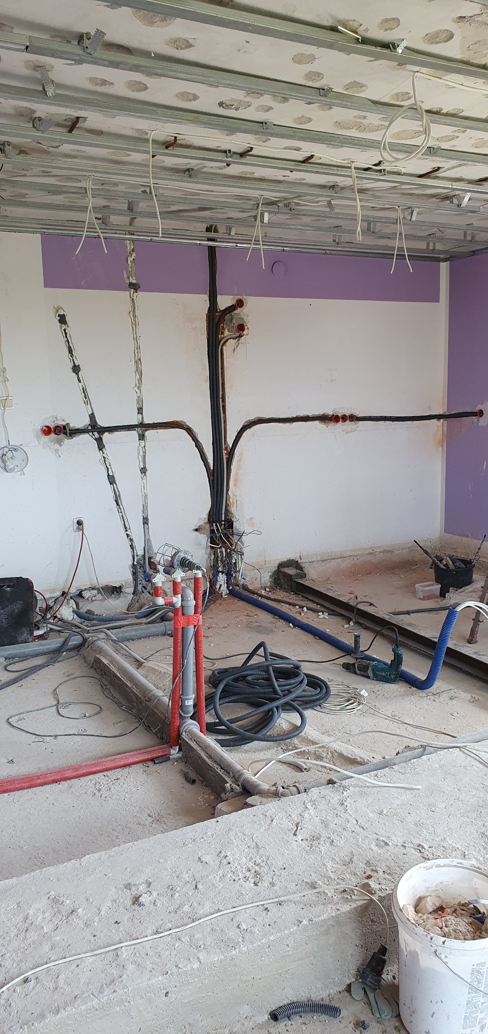 Wnętrze w trakcie remontu z widocznymi przewodami elektrycznymi i instalacjami wodno-kanalizacyjnymi, surowy beton na podłodze, częściowo pomalowane ściany na fioletowo, widoczne narzędzia...
