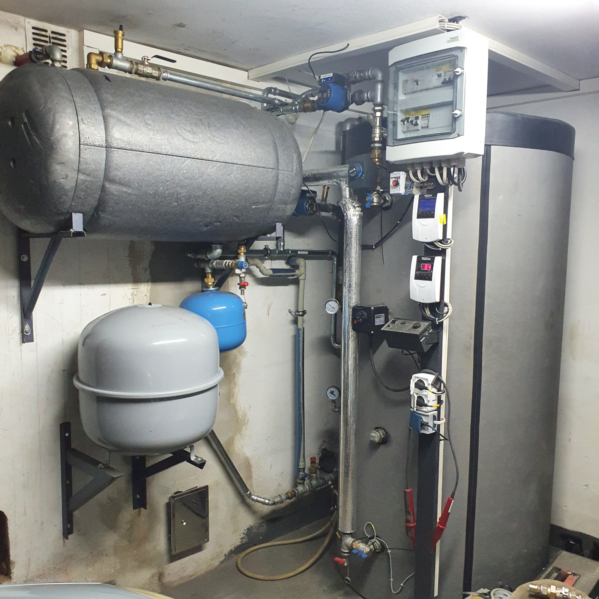 Kompleksowa instalacja grzewcza z dwoma zbiornikami, pompą obiegową i rozdzielnicą elektryczną w pomieszczeniu technicznym.