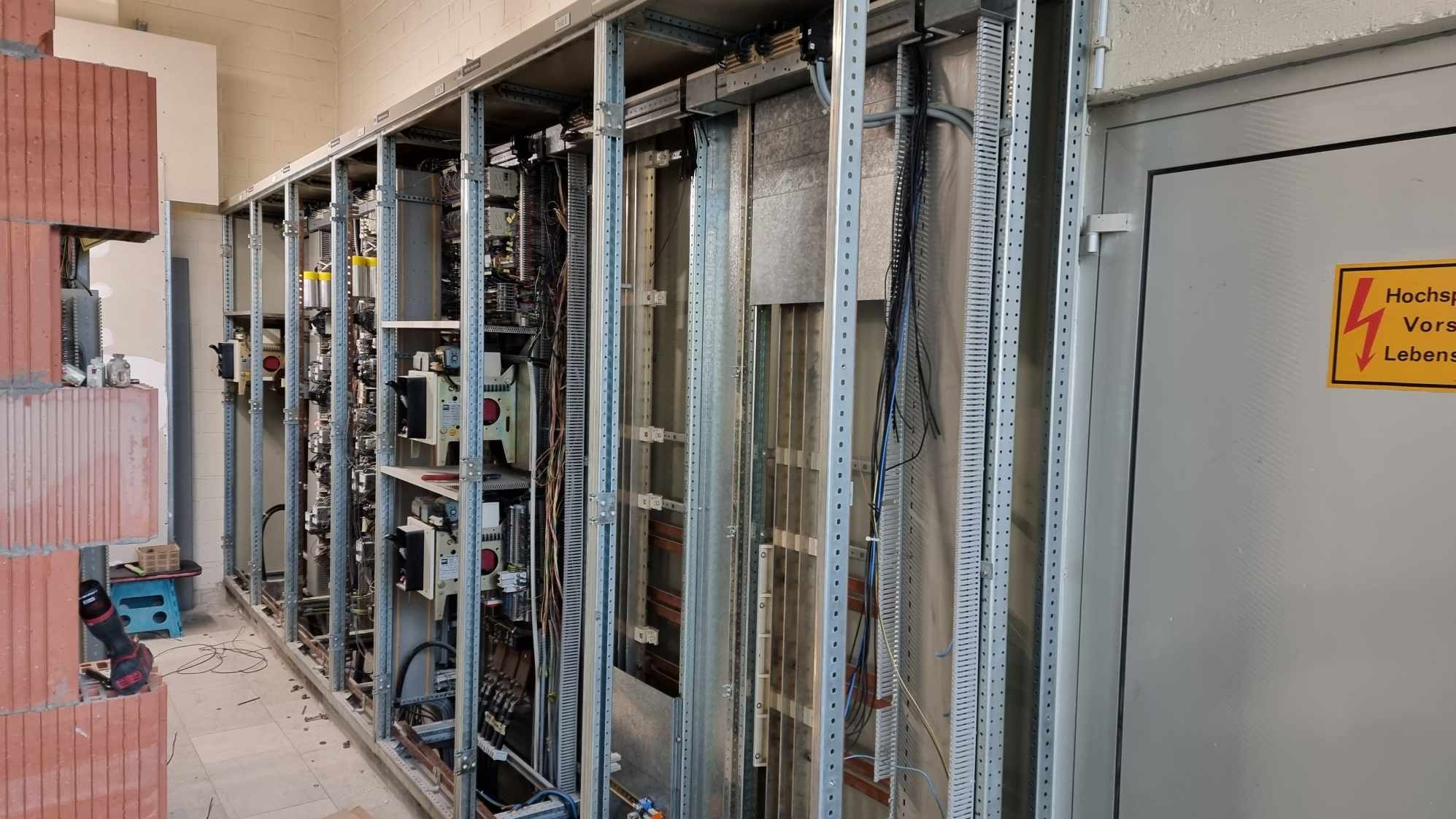 Rozbudowana szafa elektryczna w trakcie montażu, widoczne liczne kable, elementy sterujące i metalowa konstrukcja nośna, obok ściana z cegieł i tabliczka ostrzegawcza.
