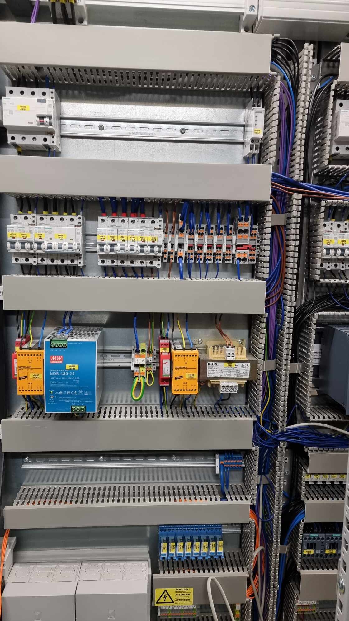 Wnętrze szarej szafy sterowniczej z widocznymi bezpiecznikami, transformatorami, zasilaczem Mean Well NDR-480-24, oraz licznymi przewodami w różnych kolorach, ułożonymi w korytkach kablowych.
