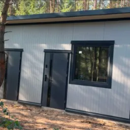 Gotowa hala z płyt warstwowych z ciemnymi oknami i drzwiami, otoczona lasem, z młodym drzewem na pierwszym planie.