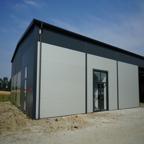 Nowoczesna hala przemysłowa z szarych płyt warstwowych, widok z zewnątrz w słoneczny dzień, widoczne okna i drzwi z ciemnymi ramami.