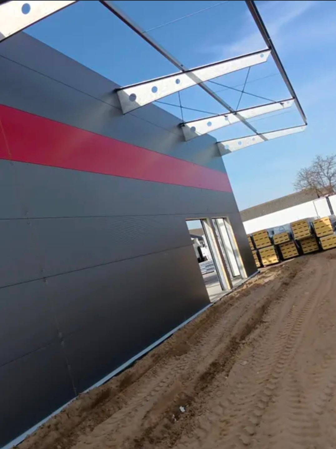 Szara hala z czerwoną linią, zadaszeniem i otwartym wejściem, widok z zewnątrz na plac budowy.