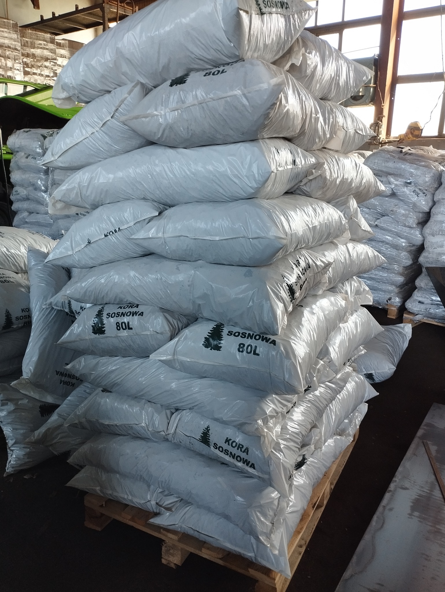 Stos worków z korą sosnową 80L ułożonych na palecie w hali magazynowej, widoczne napisy 'KORA SOSNOWA' i symbol drzewka.