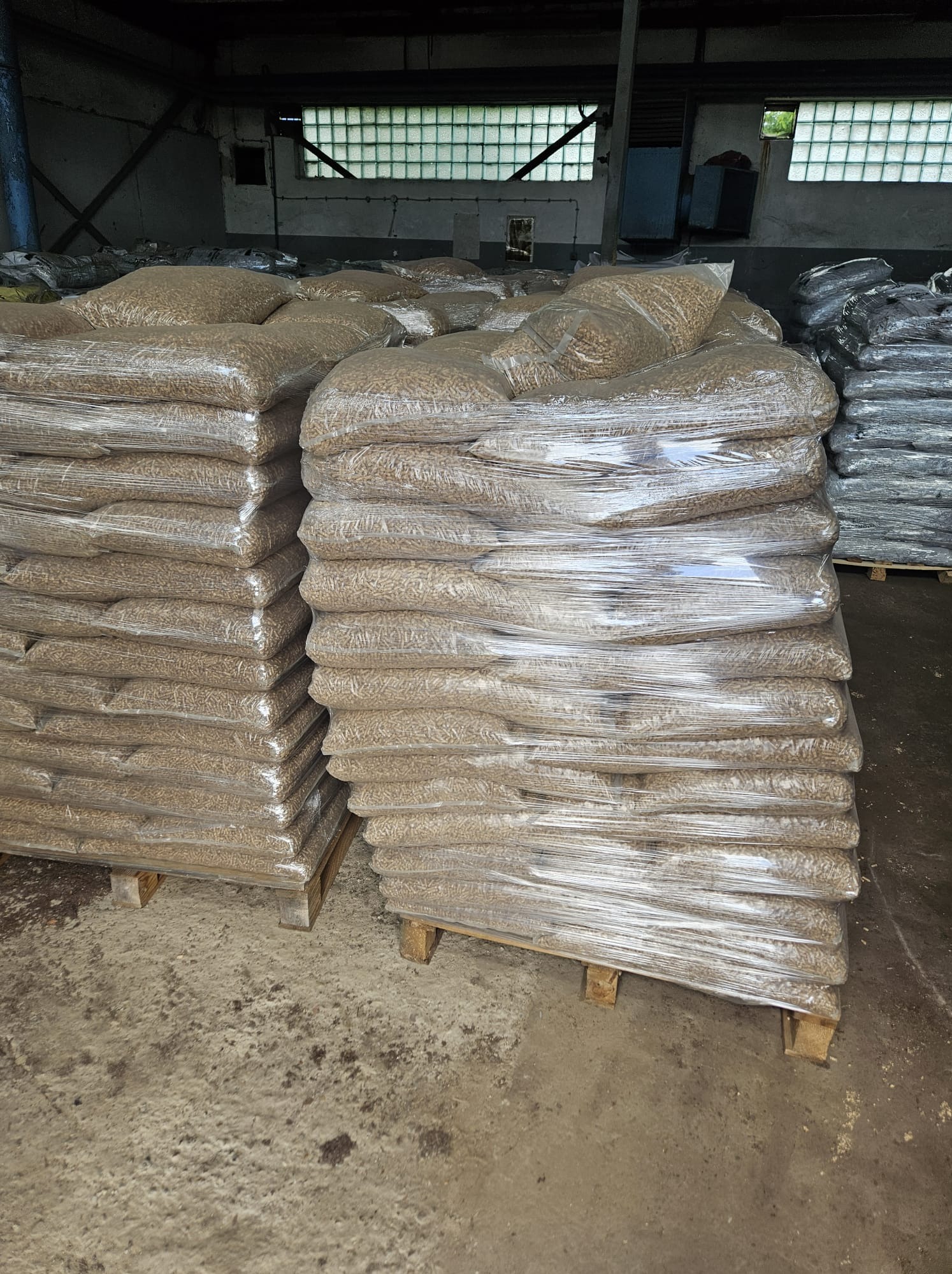 Magazyn worków z pelletem drzewnym, ułożonych na paletach i owiniętych folią stretch, widoczne okna z luksferami w tle.