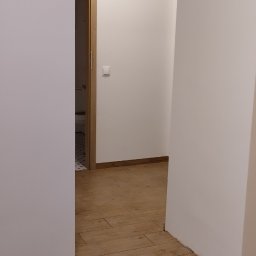 Yurii Drohomyretskyi KUB - Beżowe płytki podłogowe imitujące drewno w korytarzu z białymi ścianami. Widok na otwarte drzwi z drewnianą ościeżnicą i fragmentem podłogi w kolejnym pomieszczeniu.