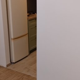 Yurii Drohomyretskyi KUB - Ukończona kuchnia z kremową lodówką i drewnopodobną podłogą. Widok na zielone szafki kuchenne w tle. Świeżo pomalowana biała ściana po prawej stronie kadru.